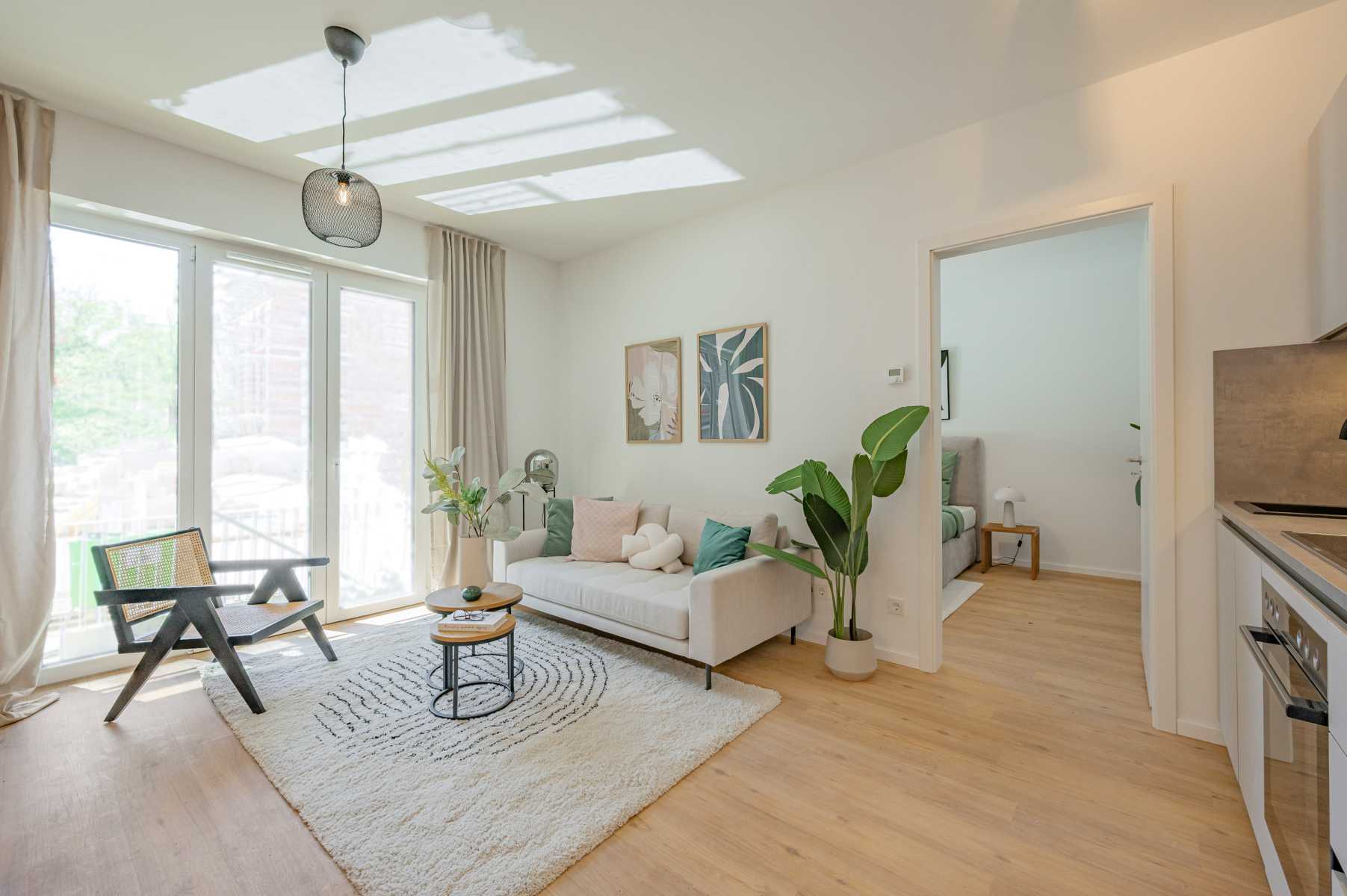Helles Wohnzimmer mit Sofa, Sessel, Pflanze und Zugang zu Schlafzimmer und Küche.