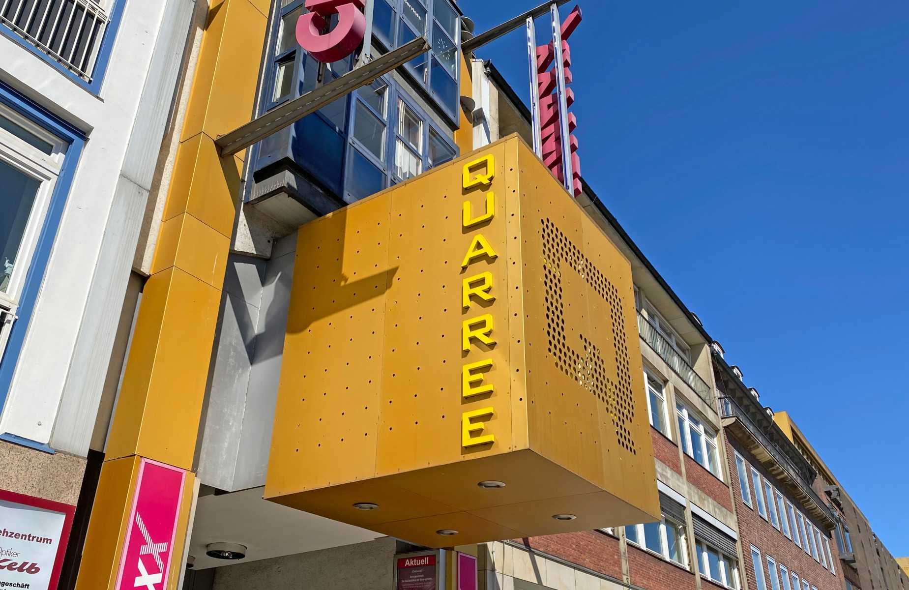 Gelbes Schild mit dem Schriftzug „QUARREE“ an einem Gebäude bei blauem Himmel.