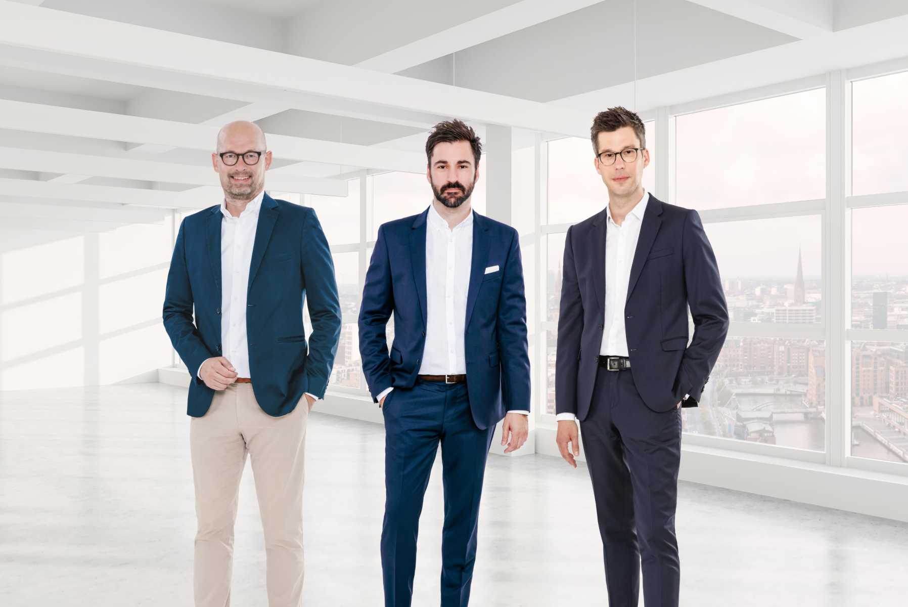 Drei Männer in Anzügen stehen in einem modernen, hellen Bürogebäude.