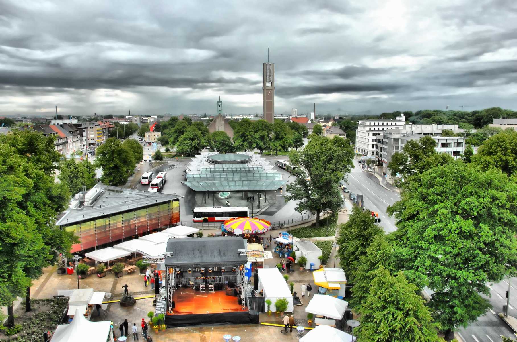 Stadtplatz mit Zelten, Bühne, Bäumen und markantem Turm unter wolkigem Himmel.